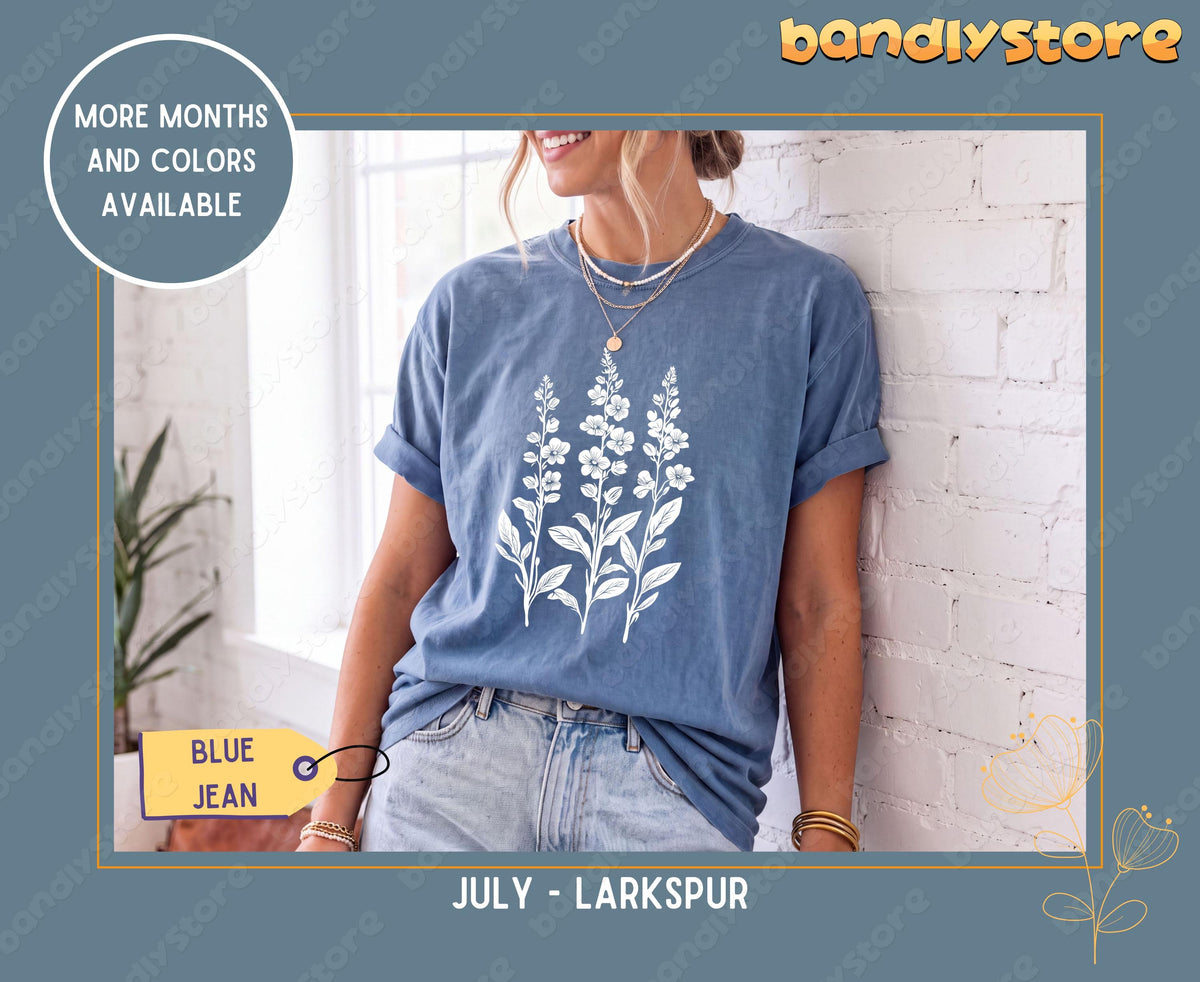 Custom Birth Month Flower T-Shirt in Blue Jean, worn by a woman leaning on a white brick wall by a window with a plant nearby.