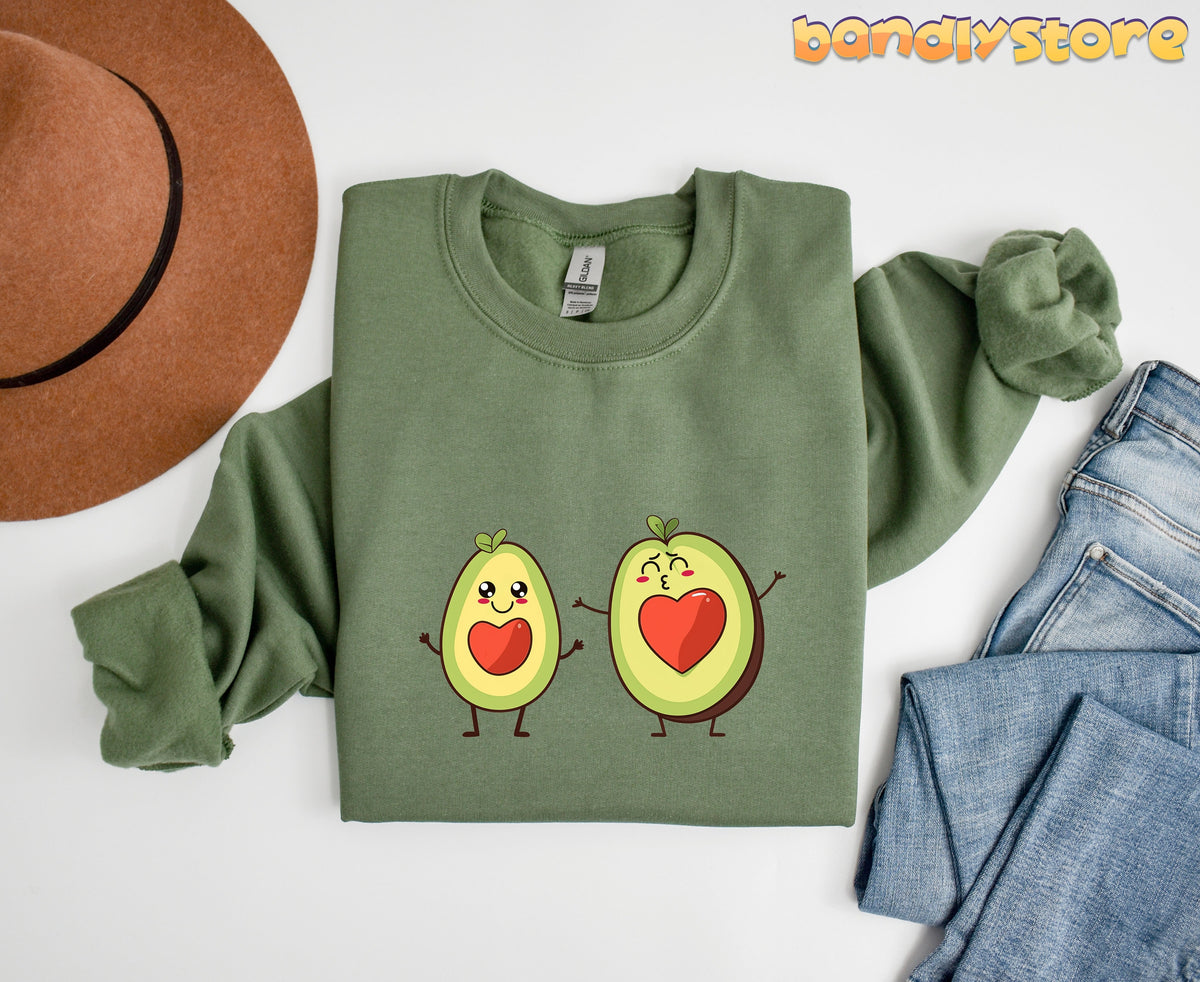 Cute Avocado Love Sweatshirt in military green, folded flat beside a brown hat and blue jeans.