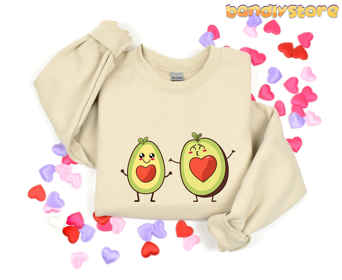 Cute Avocado Love Sweatshirt in sand laid flat with avocado pair design amid scattered heart confetti.