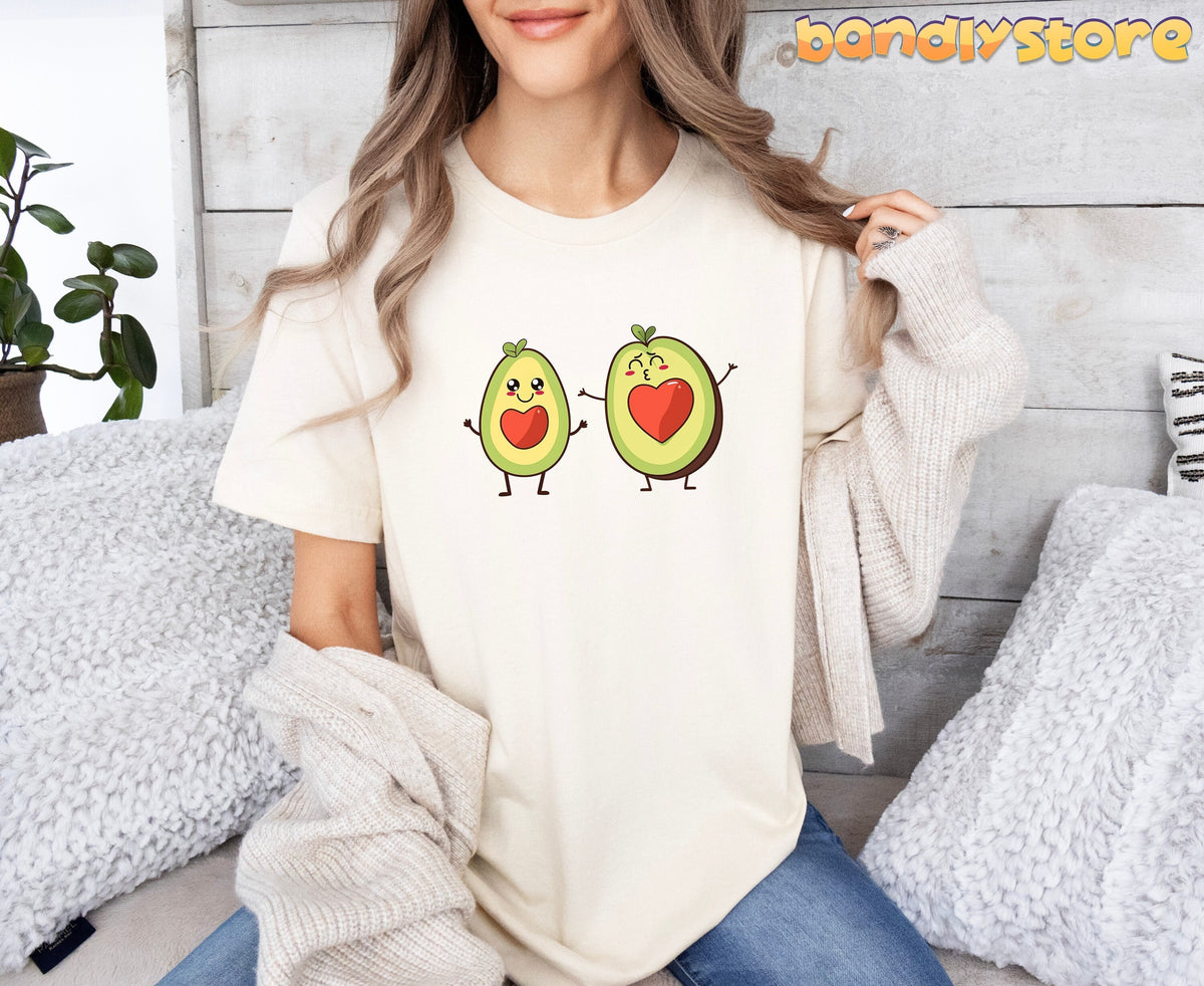 Cute Avocado Love T-Shirt in natural on a seated woman against a wooden wall with plant and pillows.