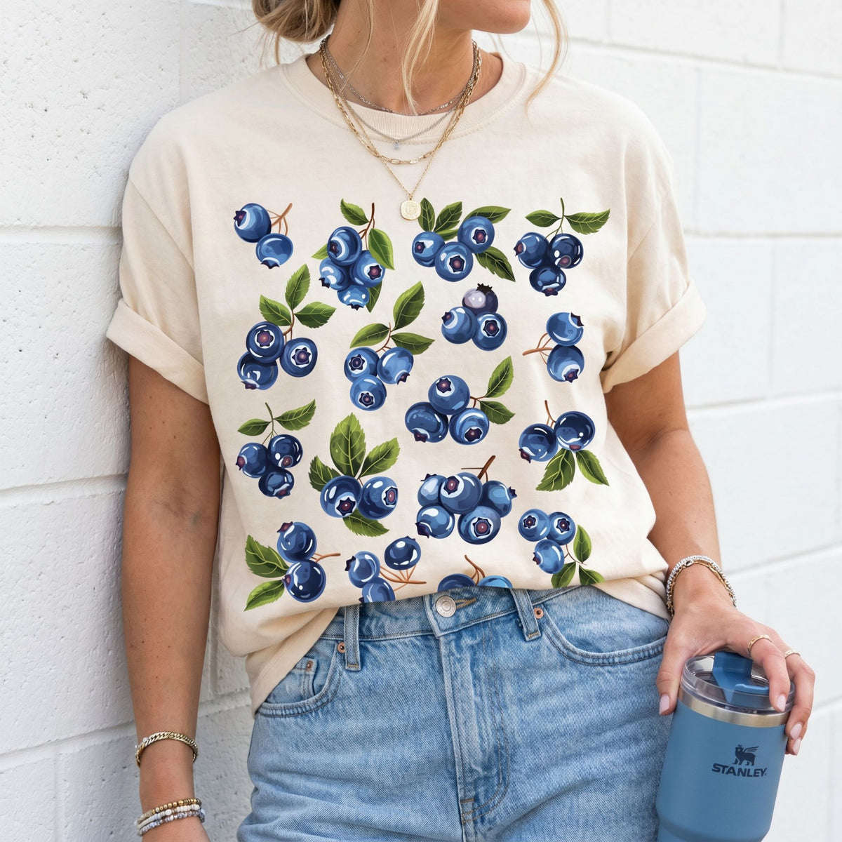 Cute Blueberry T-Shirt in ivory on a person leaning against a white brick wall holding a tumbler.