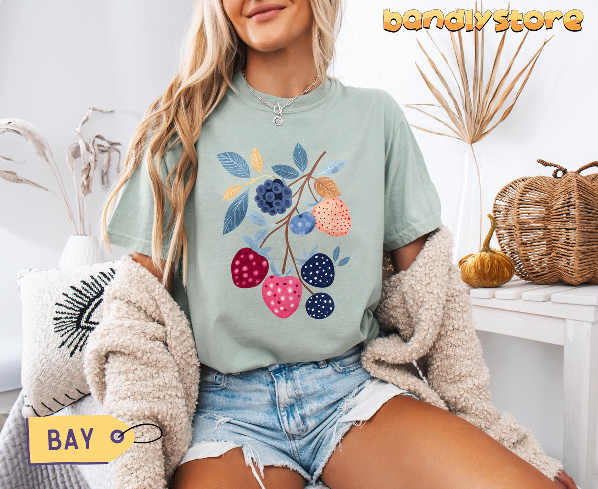 Cute Boho Strawberry T-Shirt in Bay on a seated woman indoors beside dried plants, pillow and woven basket.