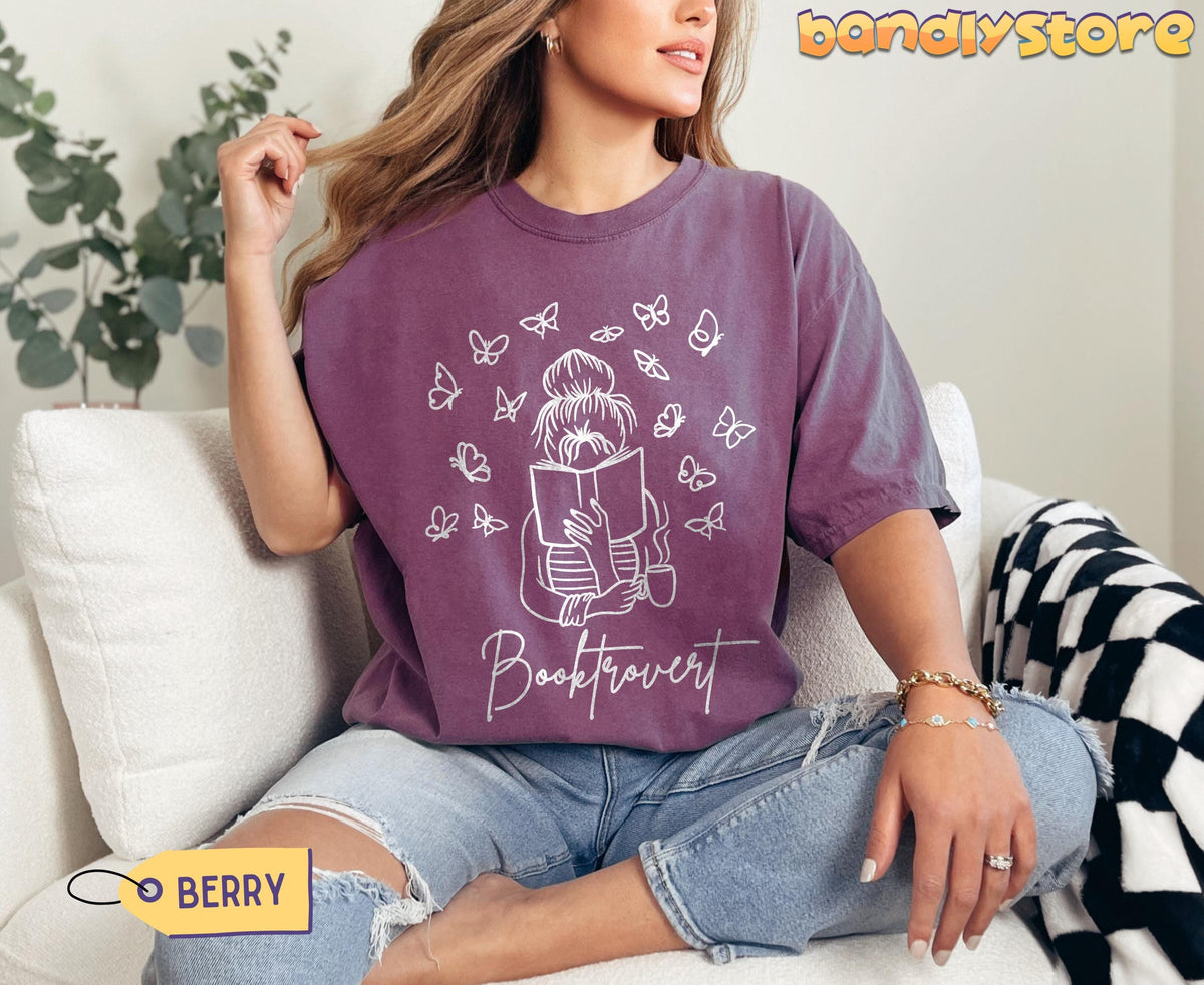 Booktrovert T-Shirt in berry on a woman seated on a white sofa by a plant and checkered blanket.