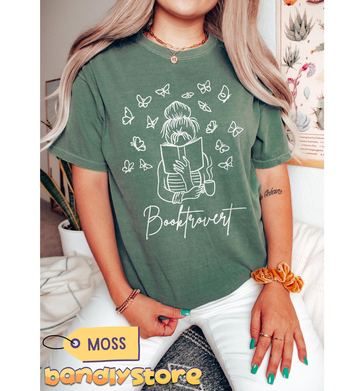 Booktrovert T-Shirt in Moss on a seated woman facing forward, indoor plants and botanical poster behind.