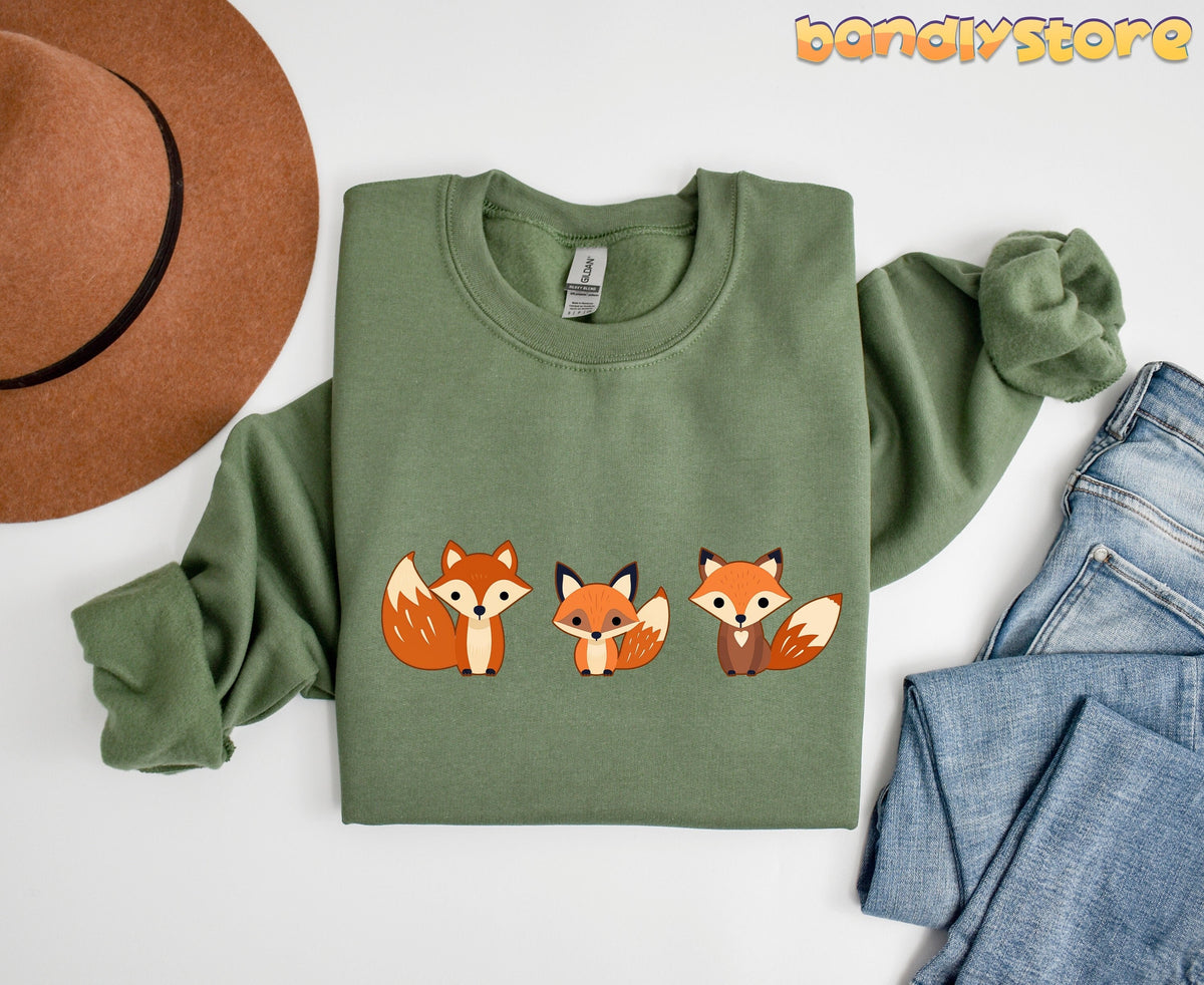 Cute Fox Sweatshirt in military green laid flat beside a brown hat and blue jeans.