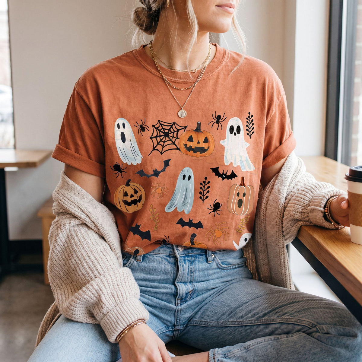 Cute Spooky Ghosts Pumpkins T-Shirt in yam on a person seated in a cafe by a window holding a coffee cup.