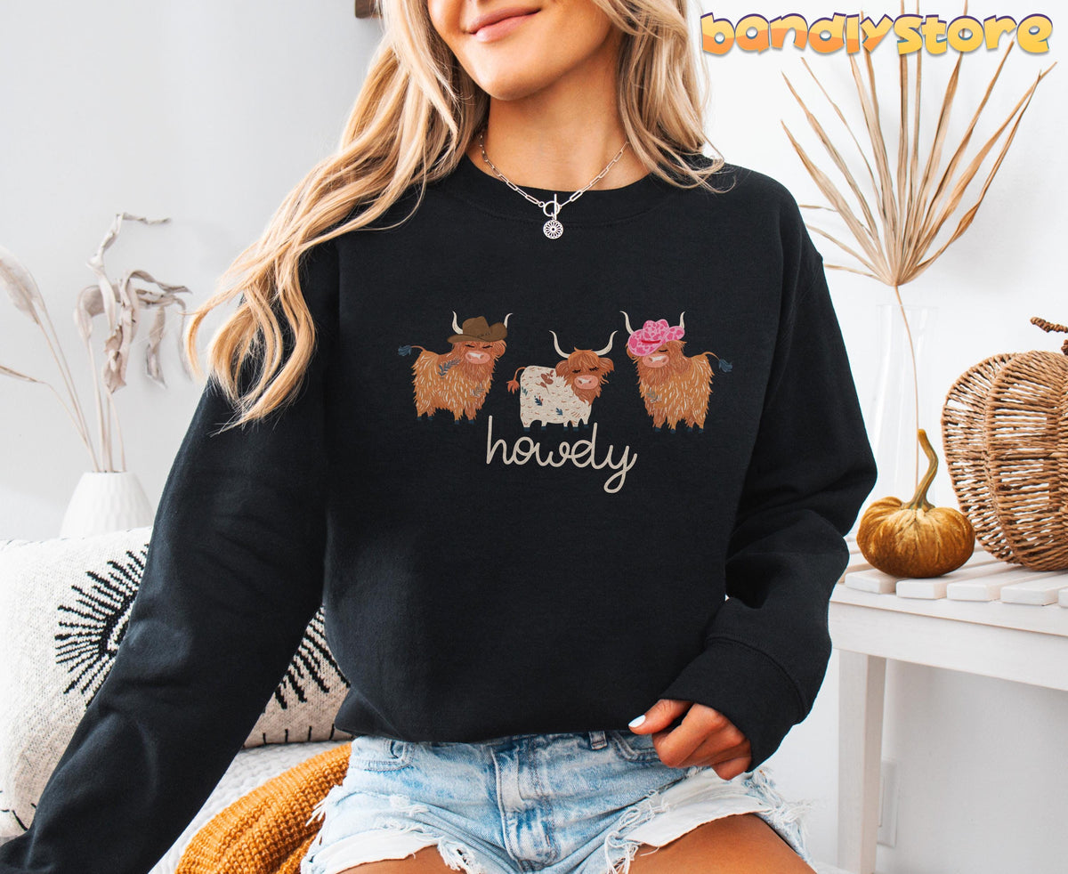 Cute Highland Cows Sweatshirt in black on a seated model amid dried plants, pumpkin and basket.