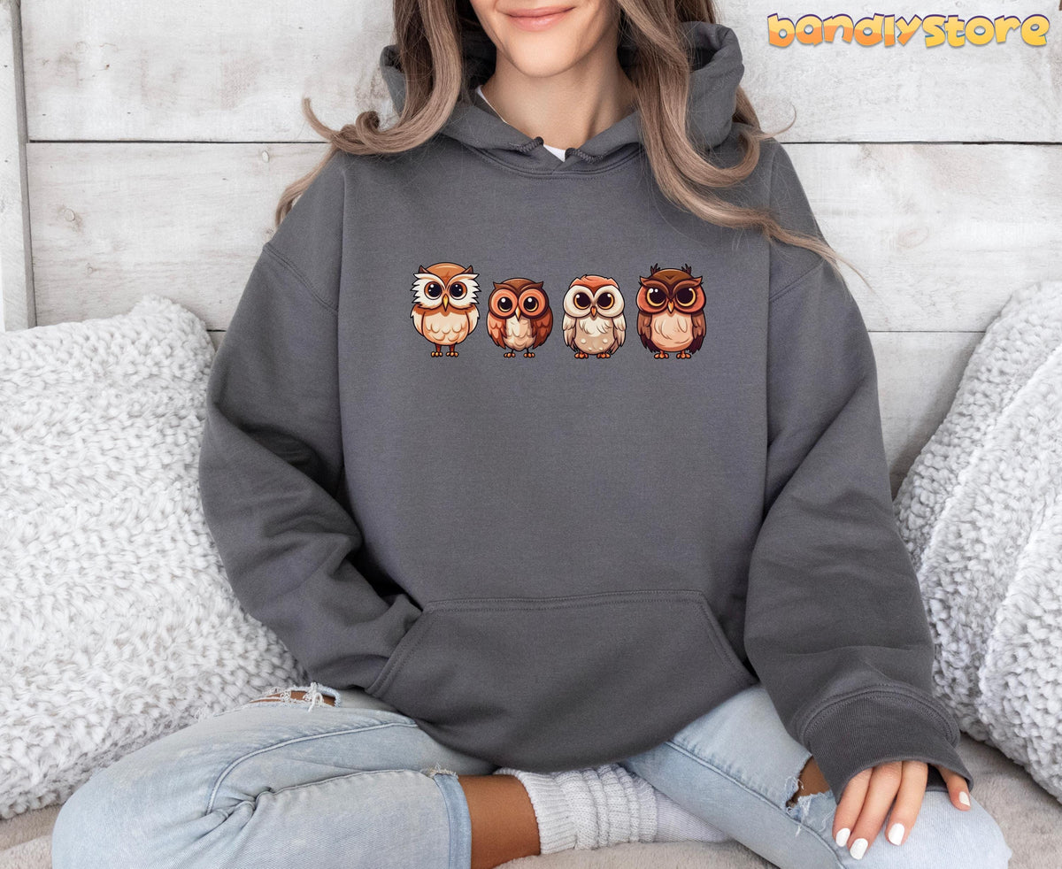 Cute Owl Hoodie in charcoal on a cross-legged person against a wooden wall flanked by white pillows.
