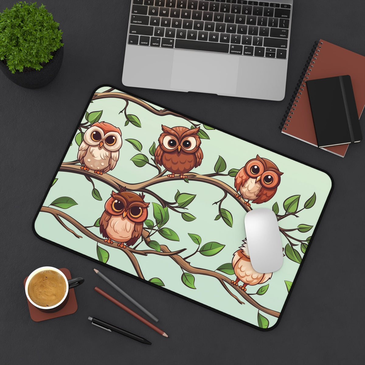 Cute Owls Desk Mat shown top-down on a dark desk with laptop, plant, coffee, pens and notebooks, 12 × 18 inches.