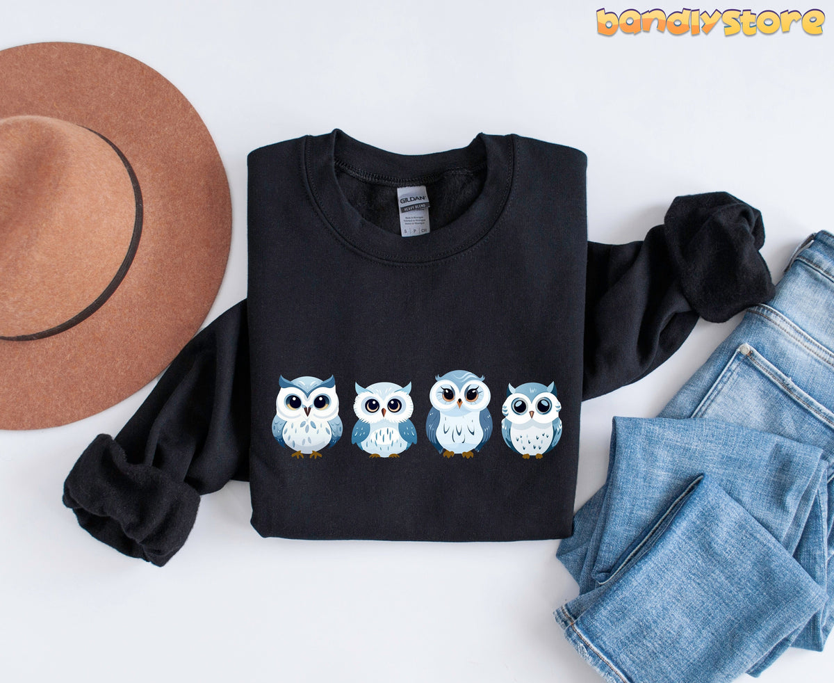 Cute Snow Owl Sweatshirt in black folded flat beside a brown hat and blue jeans.