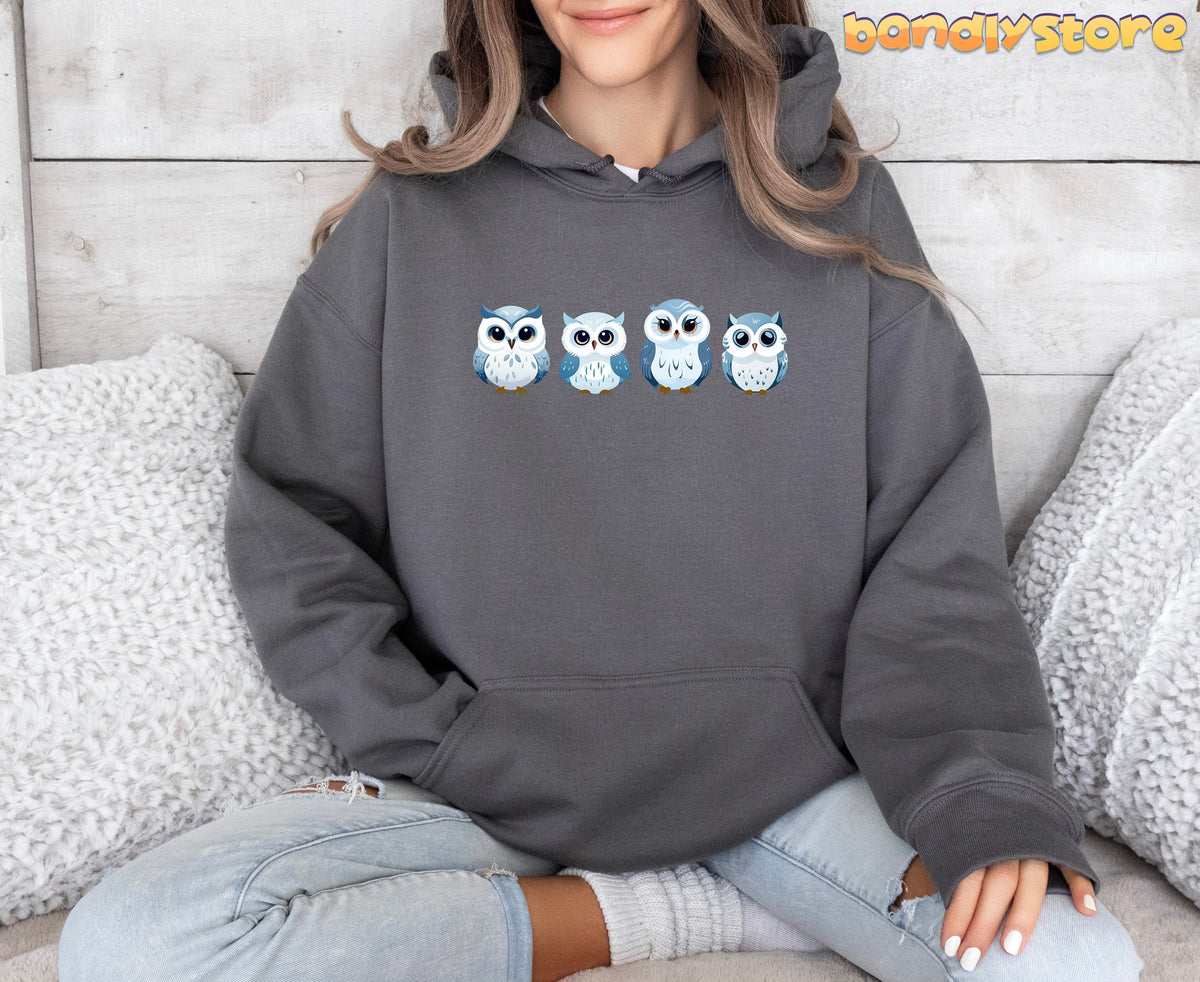 Cute Snow Owl Hoodie in charcoal, worn front-facing by a seated person amid white pillows on wood.
