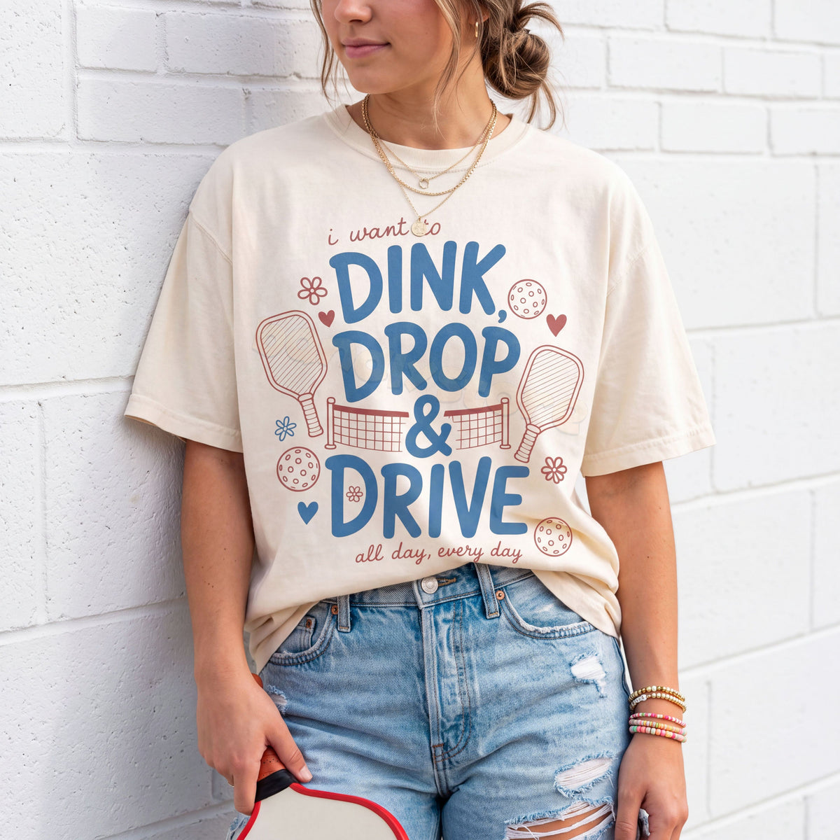 Dink Drop And Drive T-Shirt in ivory on person leaning against white brick wall holding paddle.