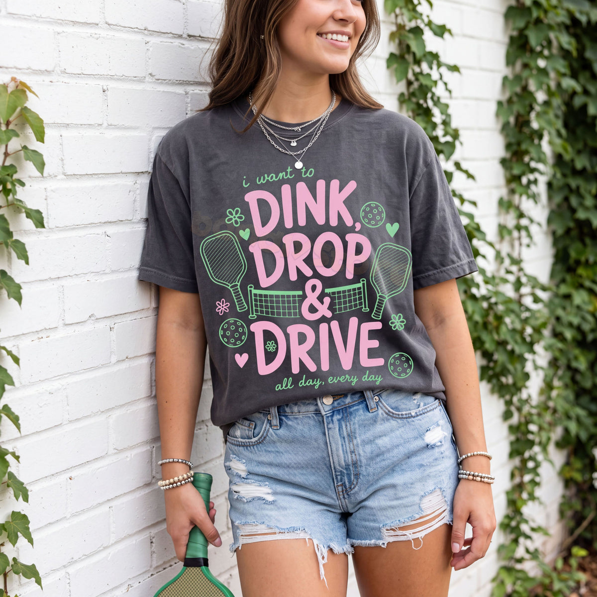 Dink Drop And Drive T-Shirt in Pepper on a woman leaning against a white brick wall with ivy, holding a paddle.