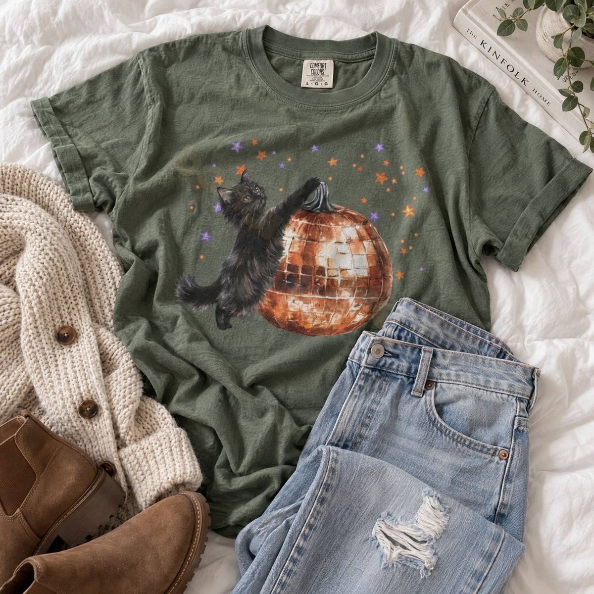 Black Cat Disco Ball Pumpkin T-Shirt in Moss, flat-lay on white bedding with jeans, boots, knit cardigan and books.