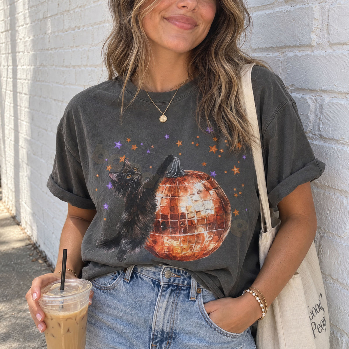 Black Cat Disco Ball Pumpkin T-Shirt in Pepper, worn outdoors by a woman holding a drink against a white brick wall.