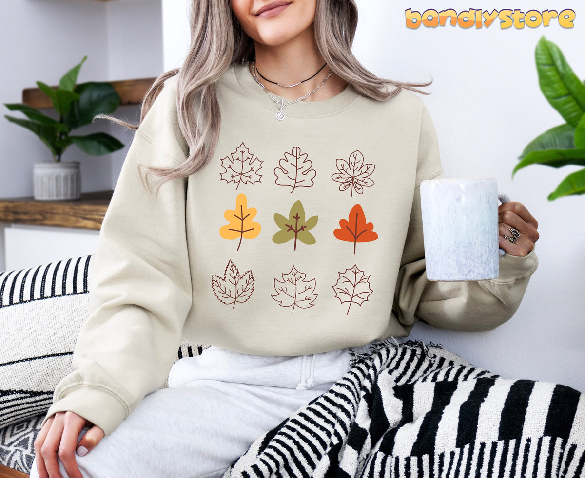 Fall Leaves Sweatshirt in Sand, worn by a seated woman holding a mug amid plants and striped throws.