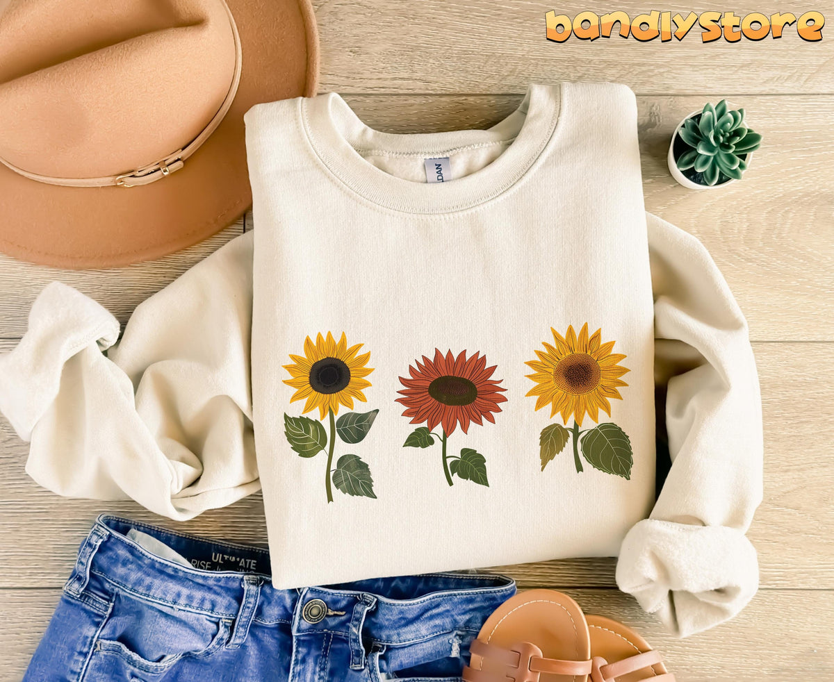 Fall Sunflower Sweatshirt in sand laid flat on wood with hat, plant, jeans and sandals.