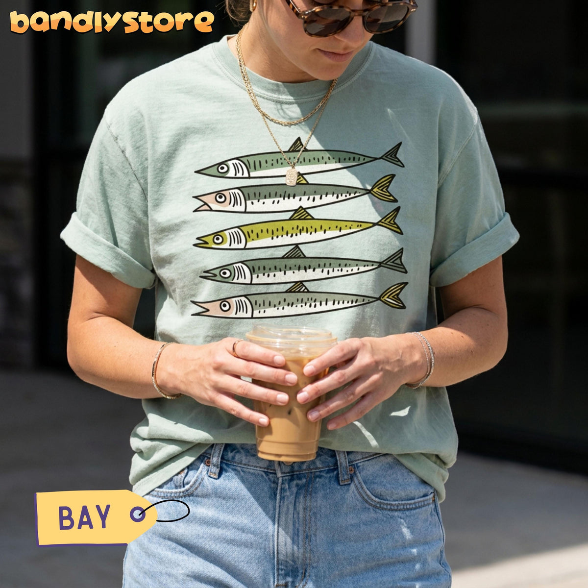 Funny Sardines T-Shirt in Bay, worn outdoors by a person holding an iced drink, five fish stacked on the chest.