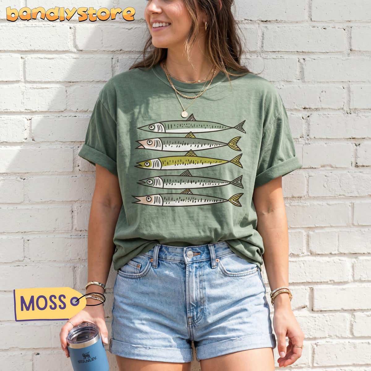 Funny Sardines T-Shirt in Moss, front view on a woman against white brick wall holding a tumbler.