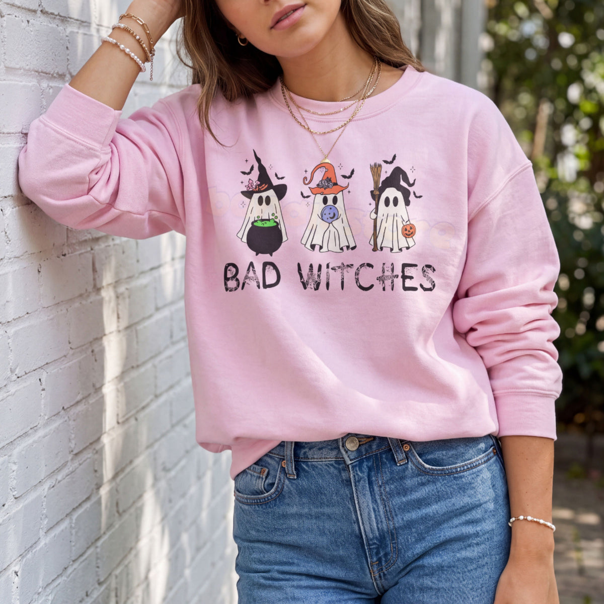 Ghost Bad Witches Sweatshirt in light pink worn by a woman leaning against a white brick wall outdoors.