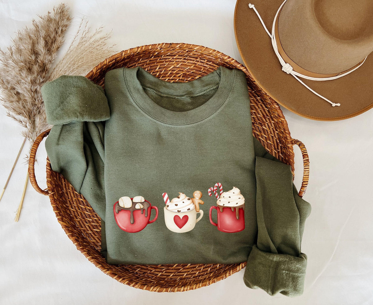 Coffee Cocoa and Gingerbread Cookies Sweatshirt in military green, folded in a woven basket beside pampas grass and a hat.
