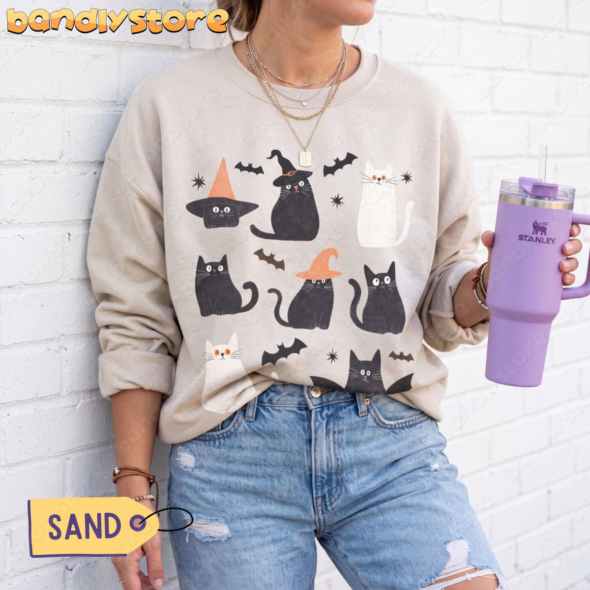Cute Halloween Black Cat Sweatshirt in sand on a person against a white brick wall holding a purple tumbler.