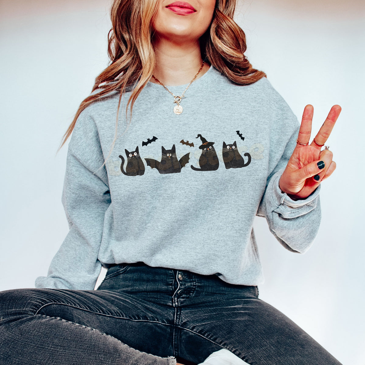 Halloween Black Cats And Bats Sweatshirt on a seated model in Sport Grey, front view with peace-sign hand and jeans.