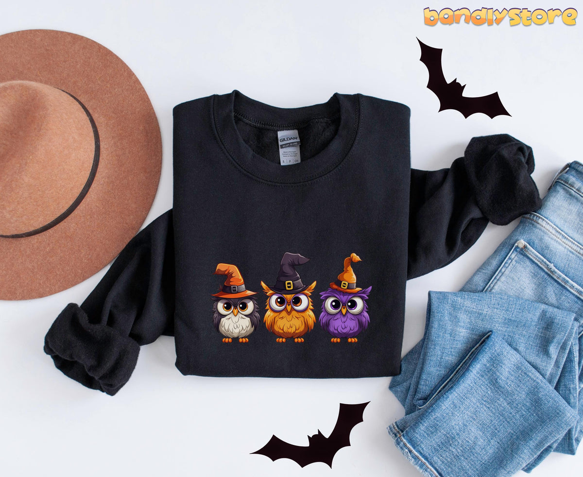 Halloween Owl Sweatshirt black folded on white surface beside a hat, jeans and bat silhouettes.