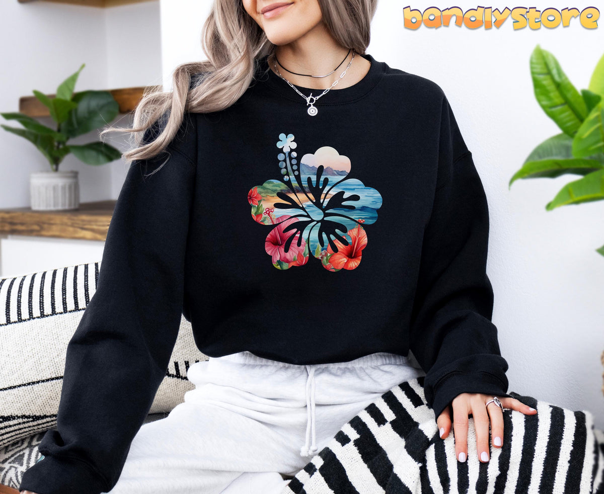Hawaii Hibiscus Sweatshirt in black on a woman seated indoors amid plants and striped cushions.