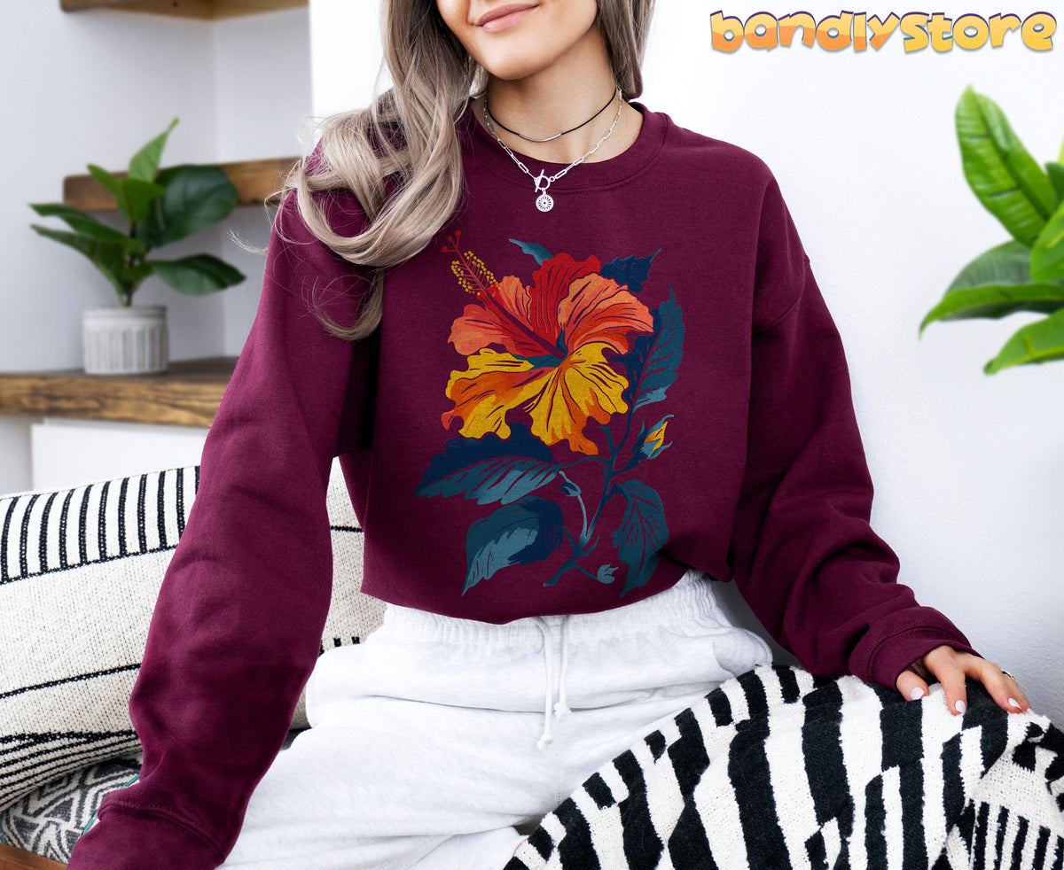 Hibiscus Flower Sweatshirt in maroon, modeled indoors amid plants, shelves, and striped textiles.