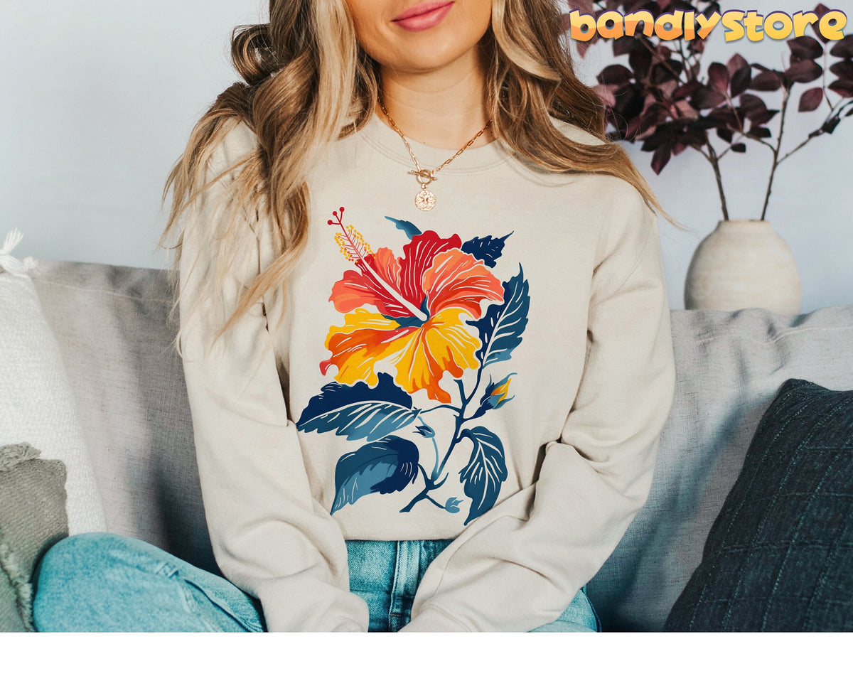 Hibiscus Flower Sweatshirt in sand on a woman seated on a sofa with pillows and a plant nearby.