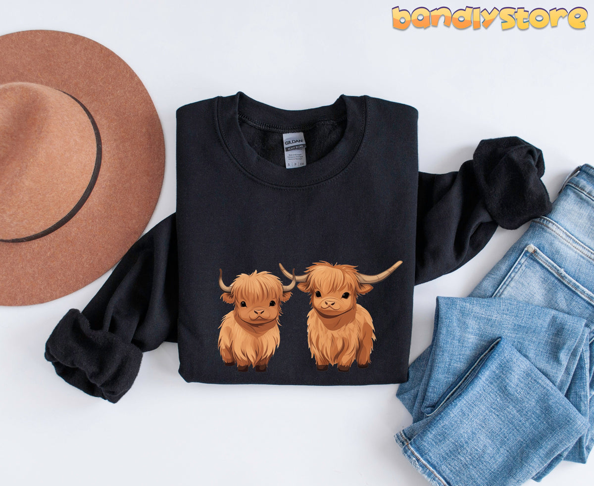 Highland Cow Sweatshirt black folded flat with hat left and jeans right on white surface.