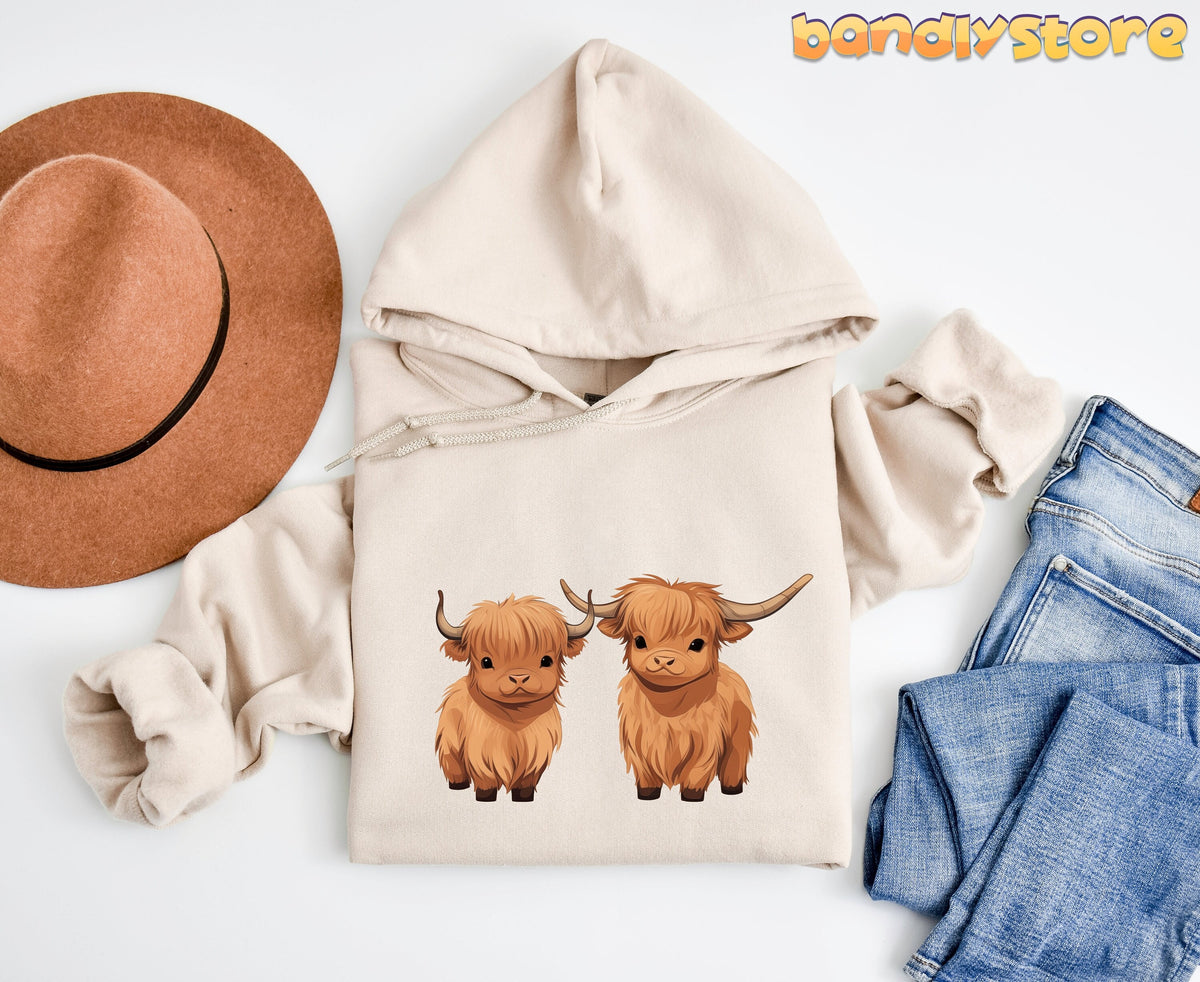 Highland Cow Hoodie in sand laid flat overhead beside a brown hat and blue jeans.