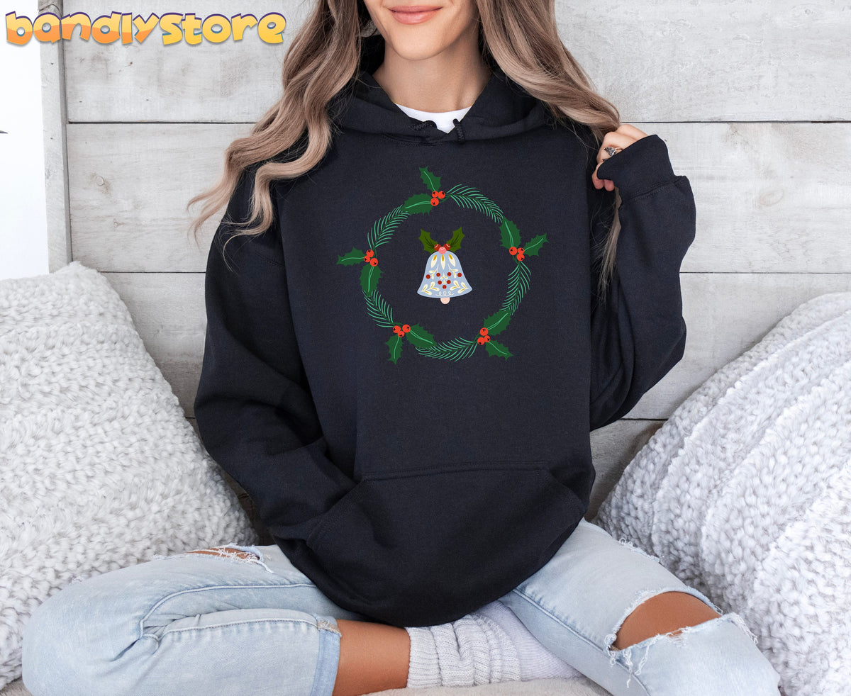 Holly Wreath Hoodie in black on a seated person by wood paneling amid white fluffy cushions.