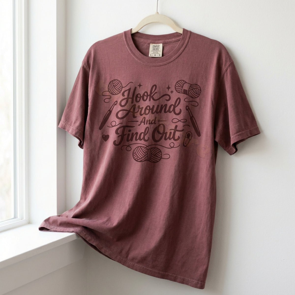 Hook Around And Find Out T-Shirt in Brick hangs on a wooden hanger by a white window, crochet design centered.