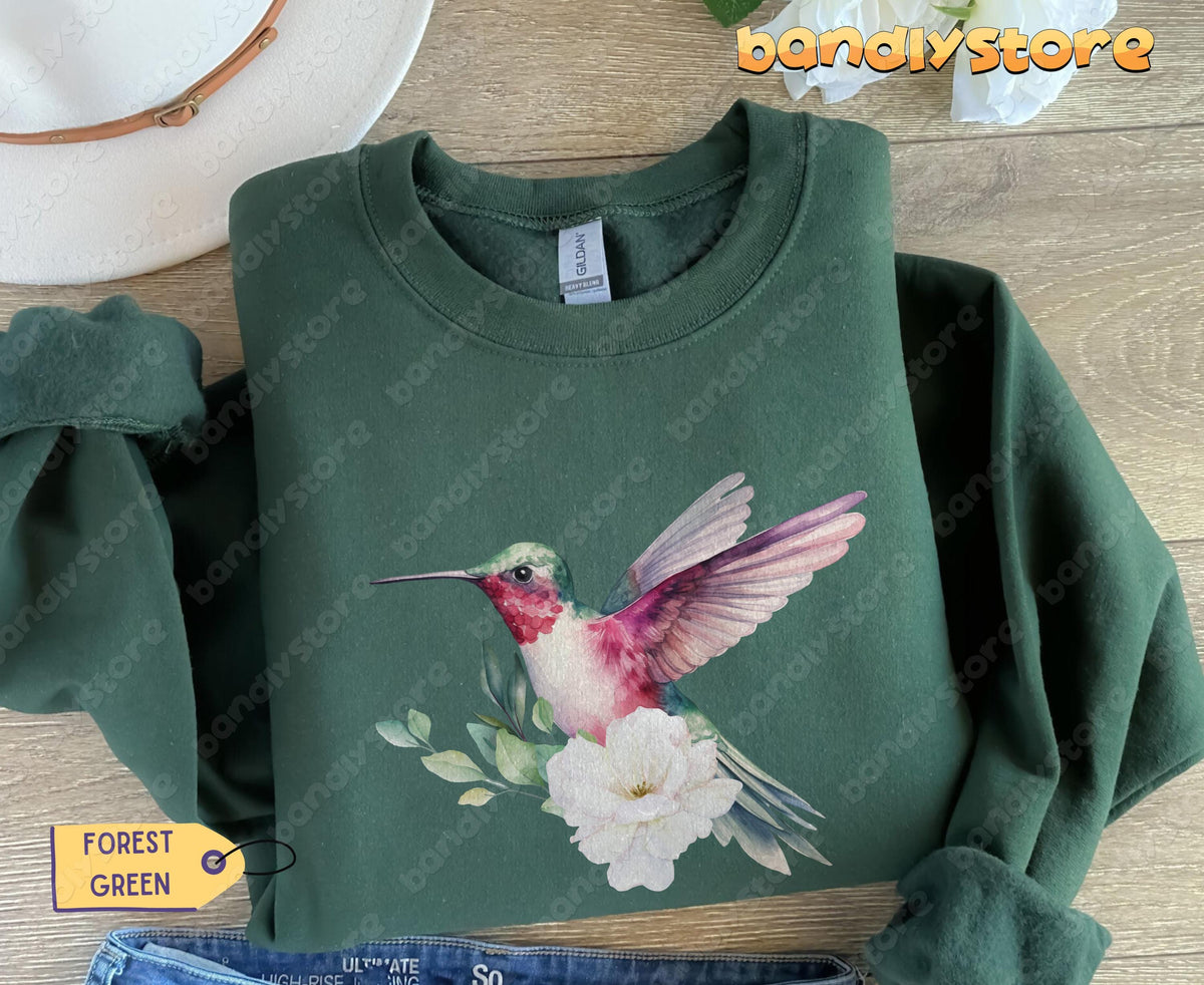 Hummingbird Flower Sweatshirt in forest green laid flat on wood beside white hat, flowers and jeans.