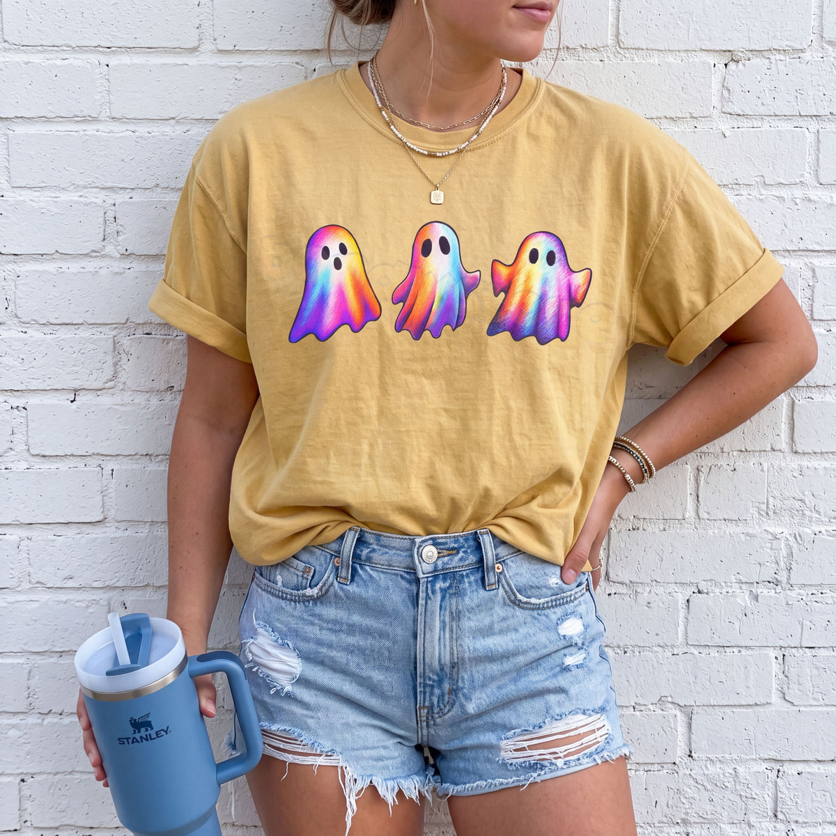 Iridescent Ghosts T-Shirt in mustard on a person facing forward against a white brick wall holding a blue tumbler.