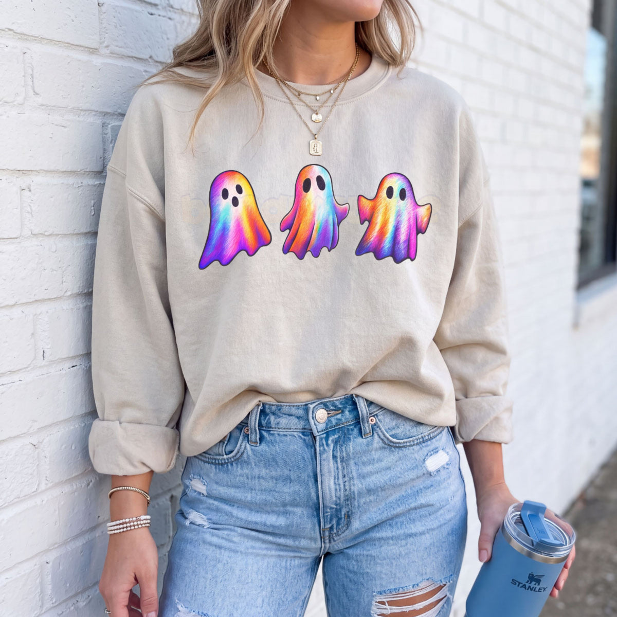 Iridescent Ghosts Sweatshirt in sand on person against white brick wall holding blue tumbler.