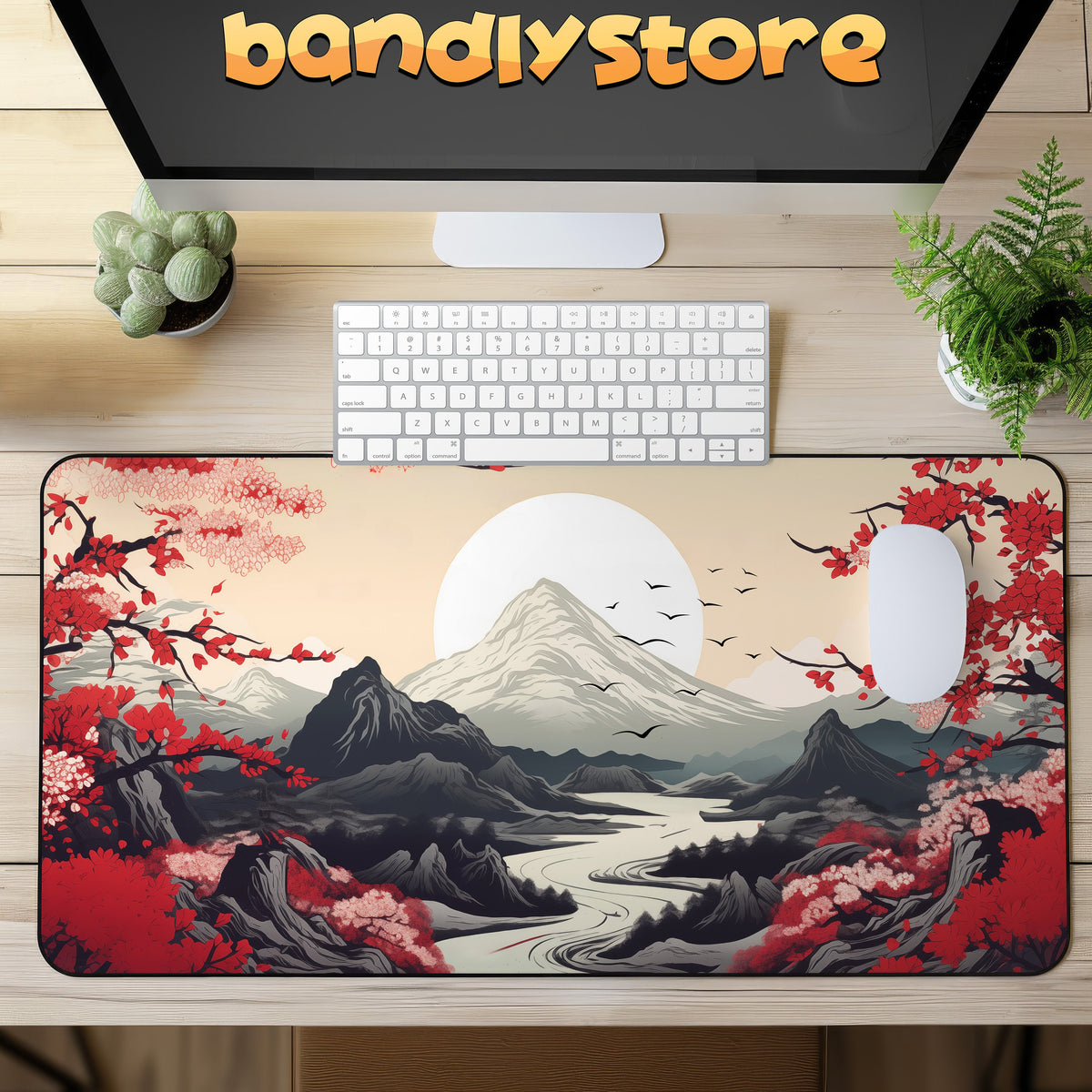 Japanese Mountains Desk Mat on wooden desk under keyboard and mouse beside monitor with bandlystore logo and plants, top view.