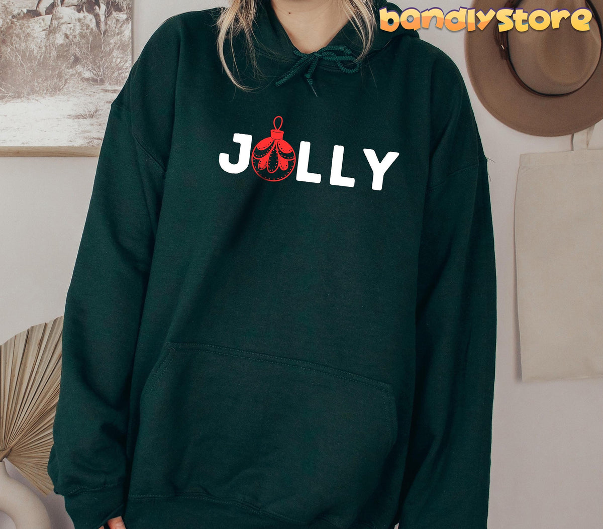 Feeling Jolly Christmas Hoodie in forest green, front view on a person amid wall art, hat and tote.