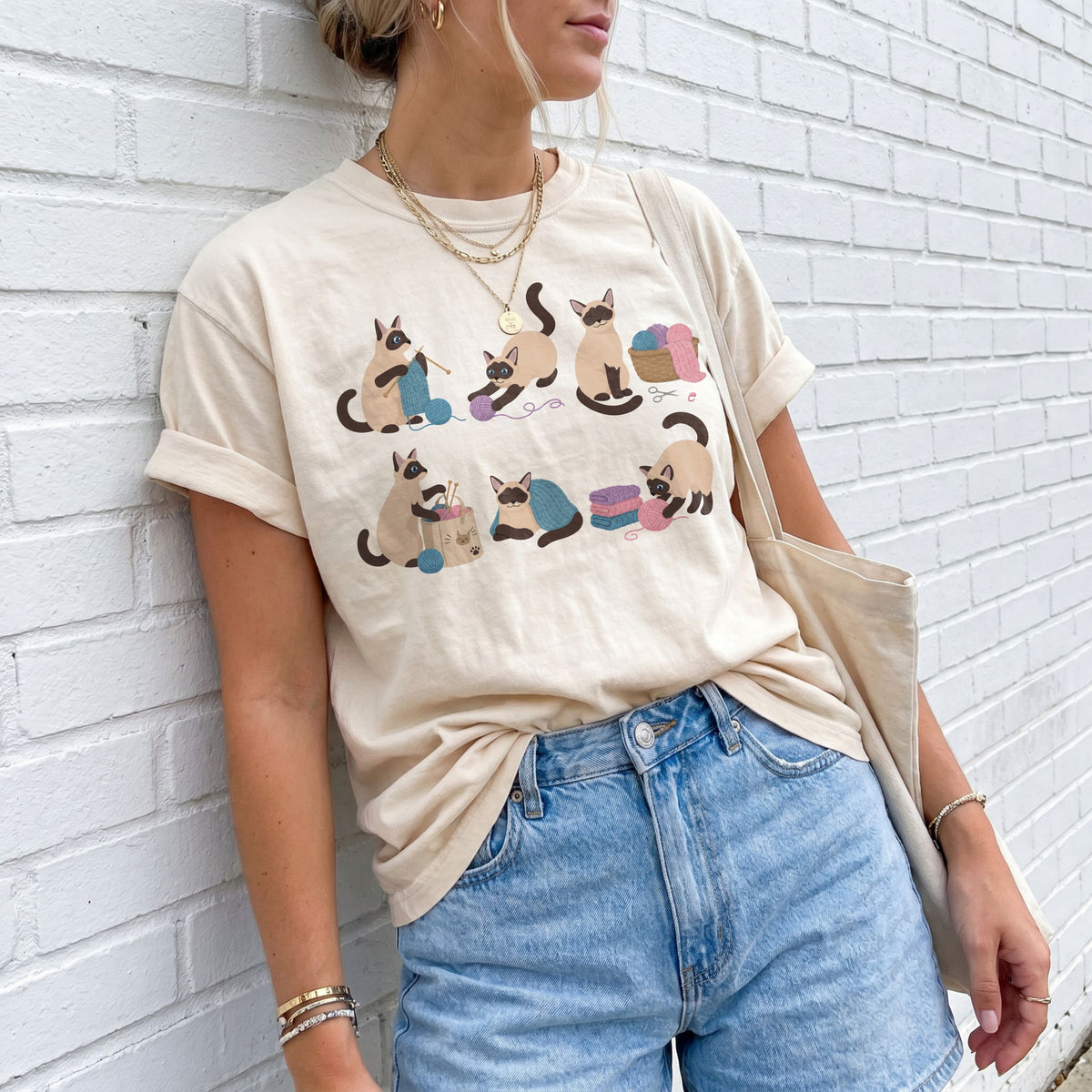 Funny Knitting Siamese Cats T-Shirt in ivory on a person leaning by a white brick wall with jeans and tote.