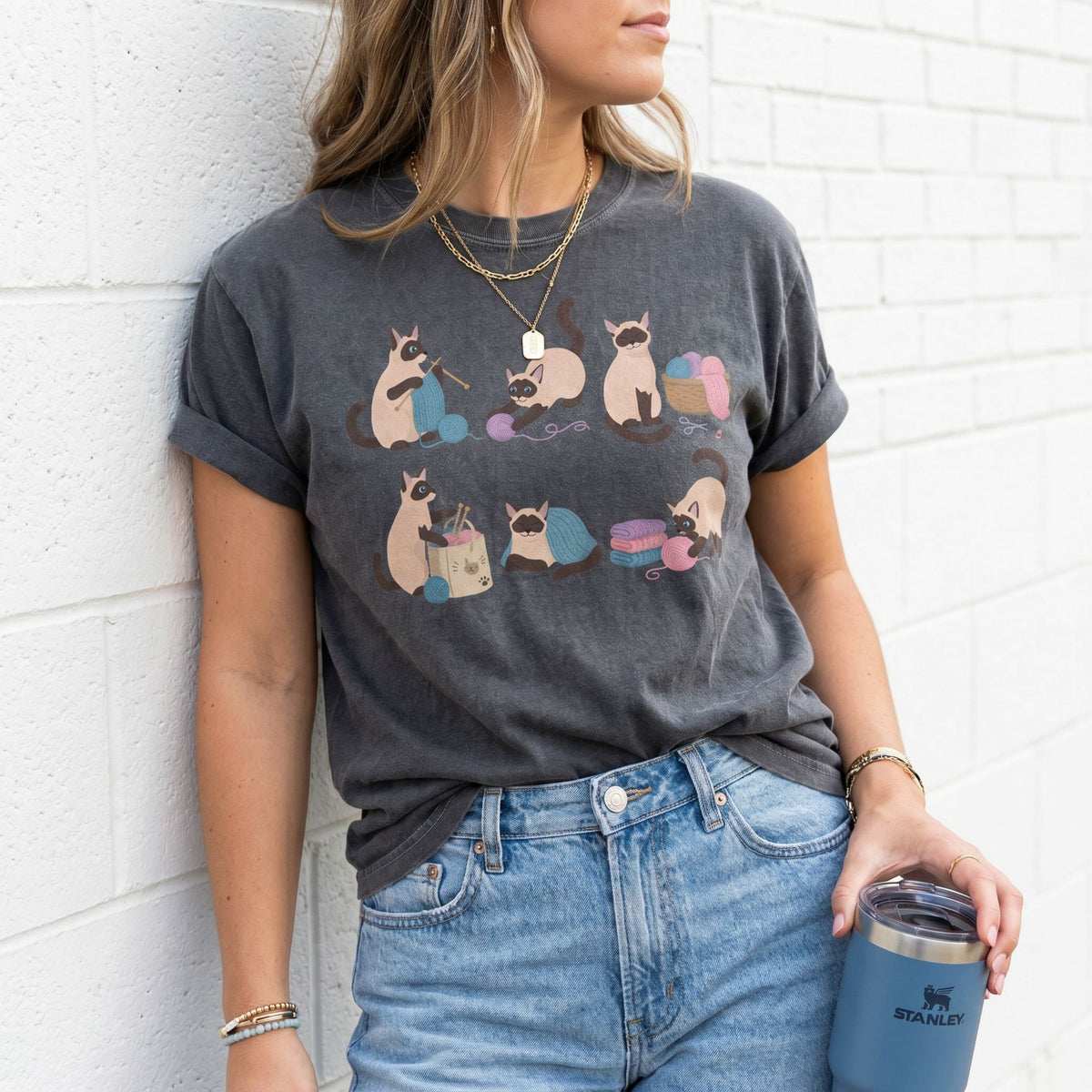 Funny Knitting Siamese Cats T-Shirt in Pepper on model leaning against white brick wall holding tumbler.