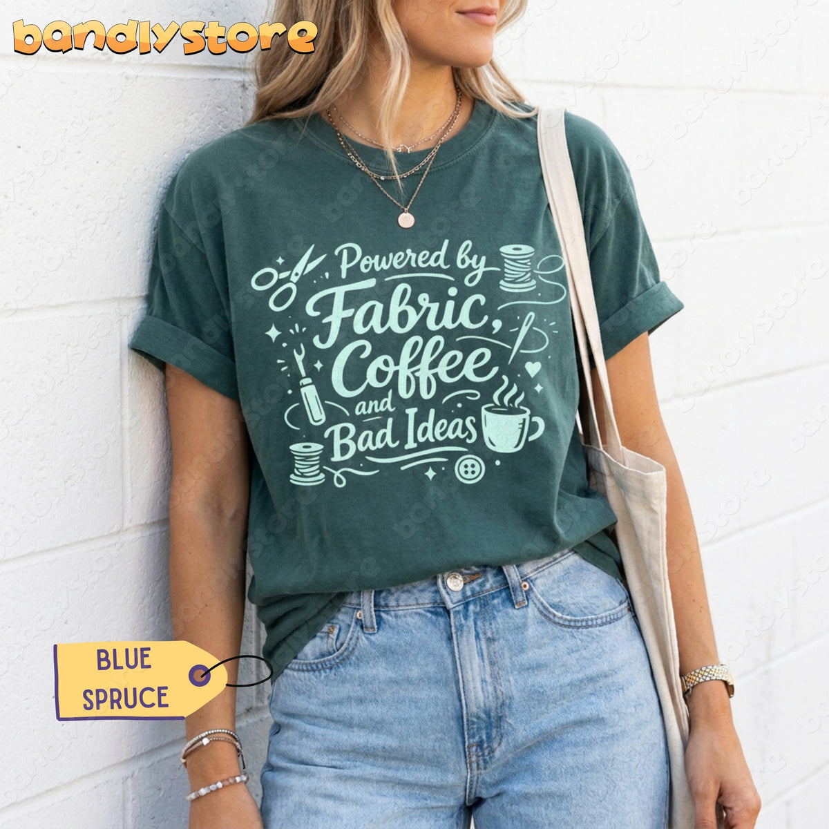 Linocut Sewing T-Shirt in Blue Spruce on a person leaning against a white brick wall with a tote bag.