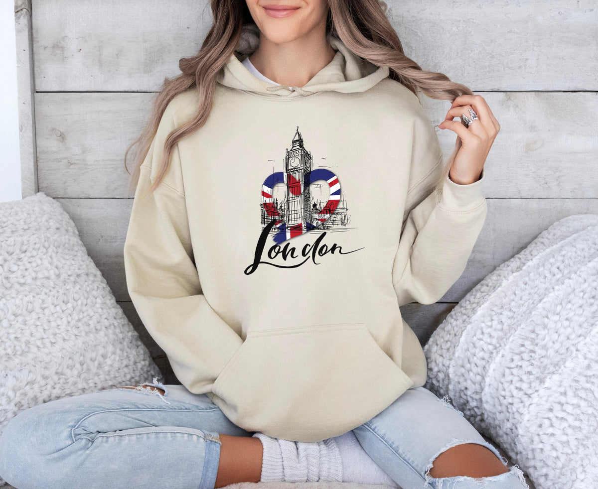 London Big Ben Hoodie in sand worn by a person sitting cross-legged amid white pillows against a wood wall.