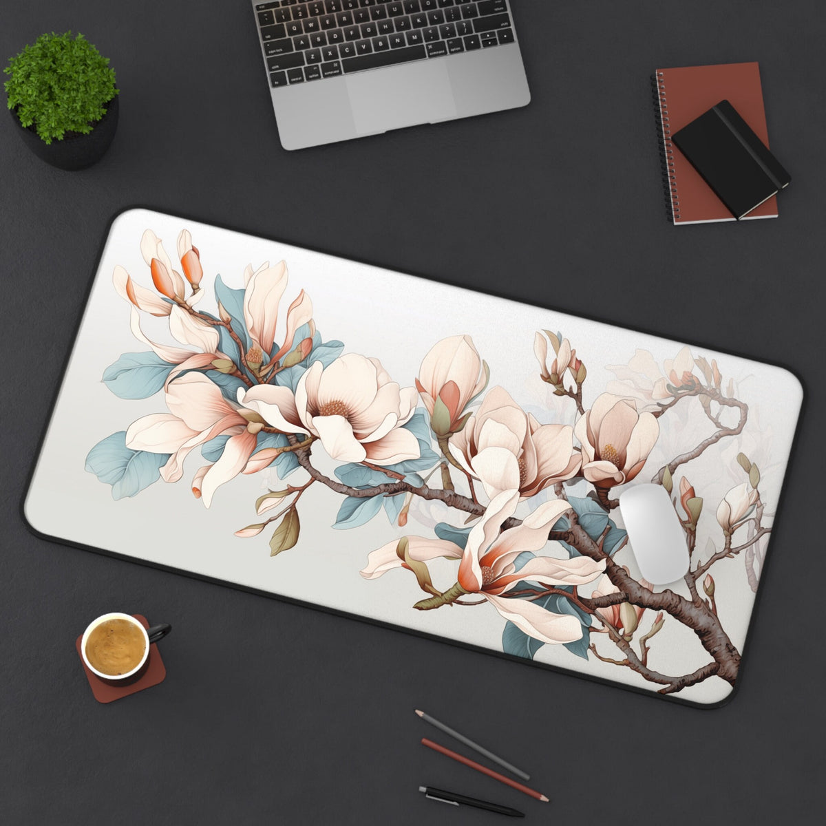 Magnolia Flowers Desk Mat 16 × 32 inches from above on a dark desk with laptop, plant, notebooks, coffee, pens and mouse.