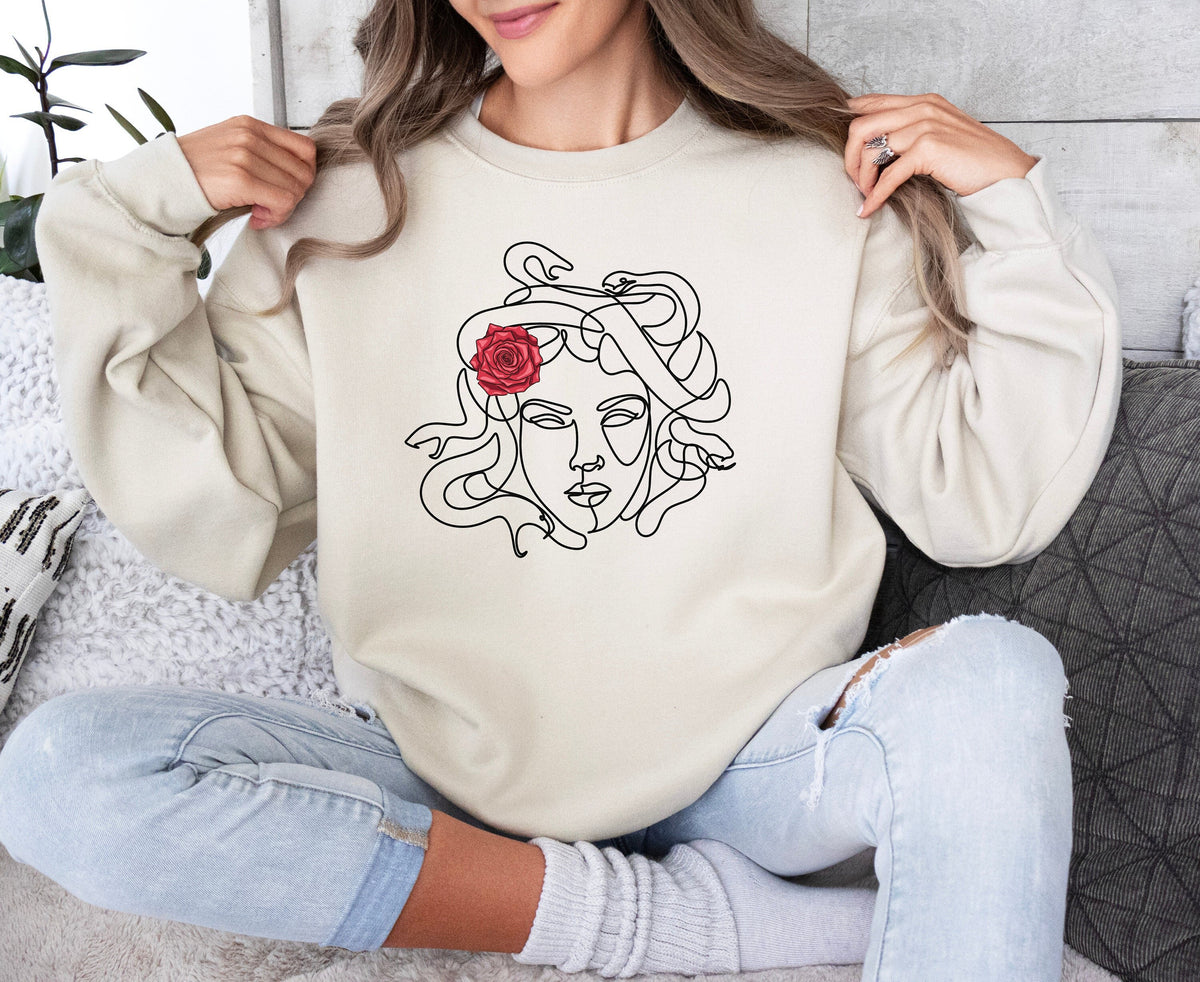 Medusa Sweatshirt in sand on seated woman holding collar, pillows and plant nearby.