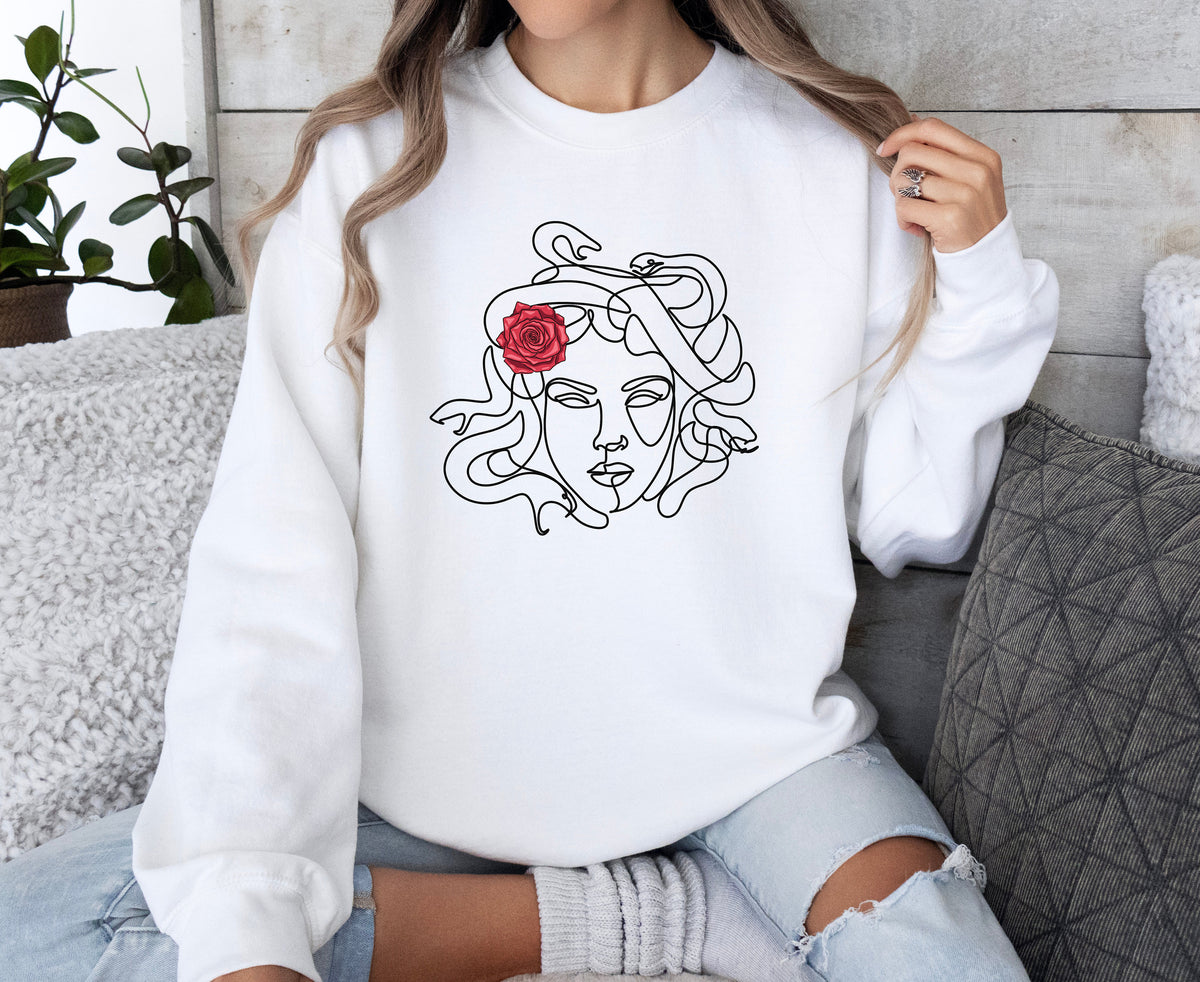 Medusa Sweatshirt in white on a person sitting cross-legged beside pillows and a plant by a wooden wall.