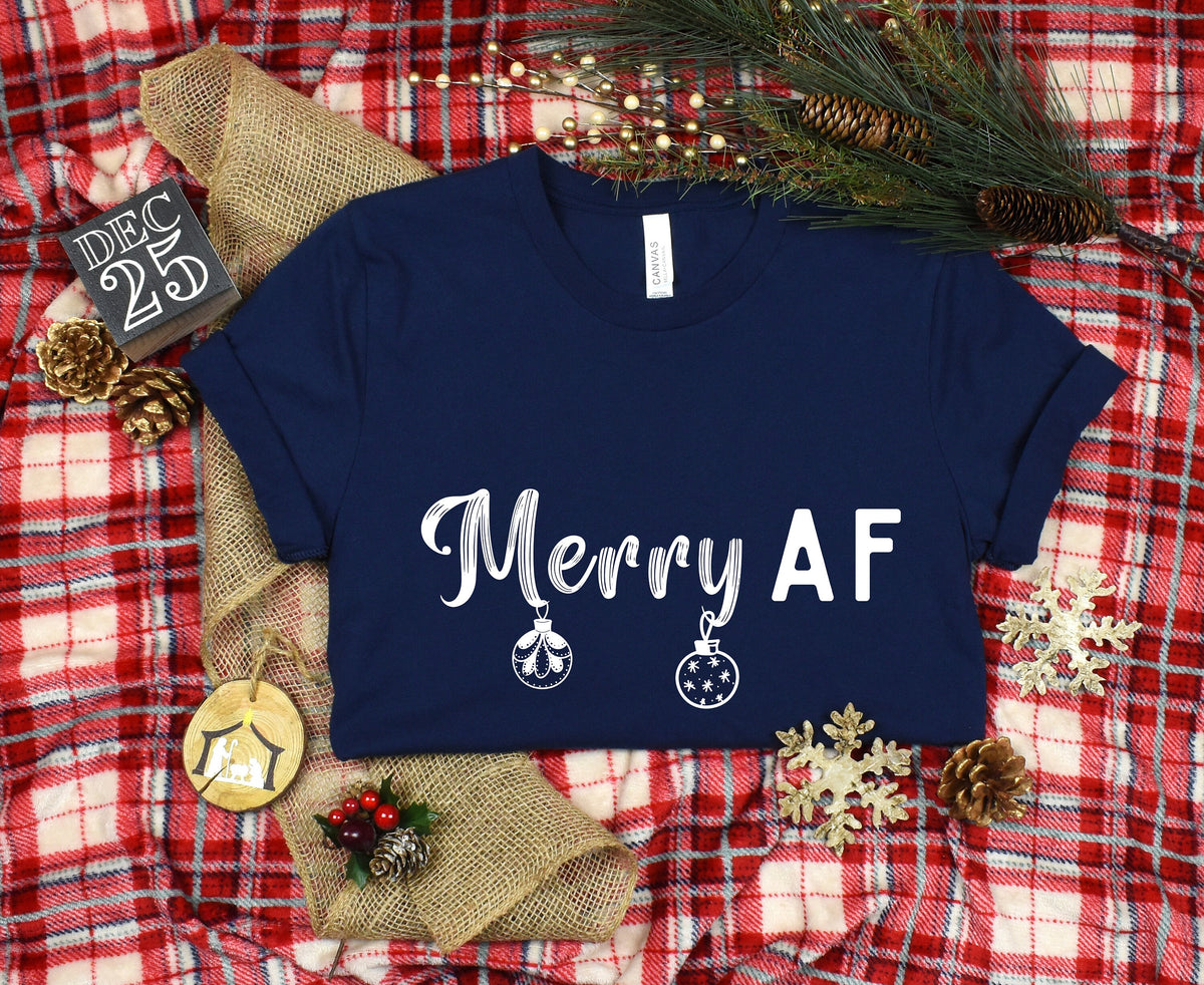 Merry AF T-Shirt in navy, flat-laid on red plaid with burlap, pine greenery, snowflakes, pinecones and a DEC 25 block.