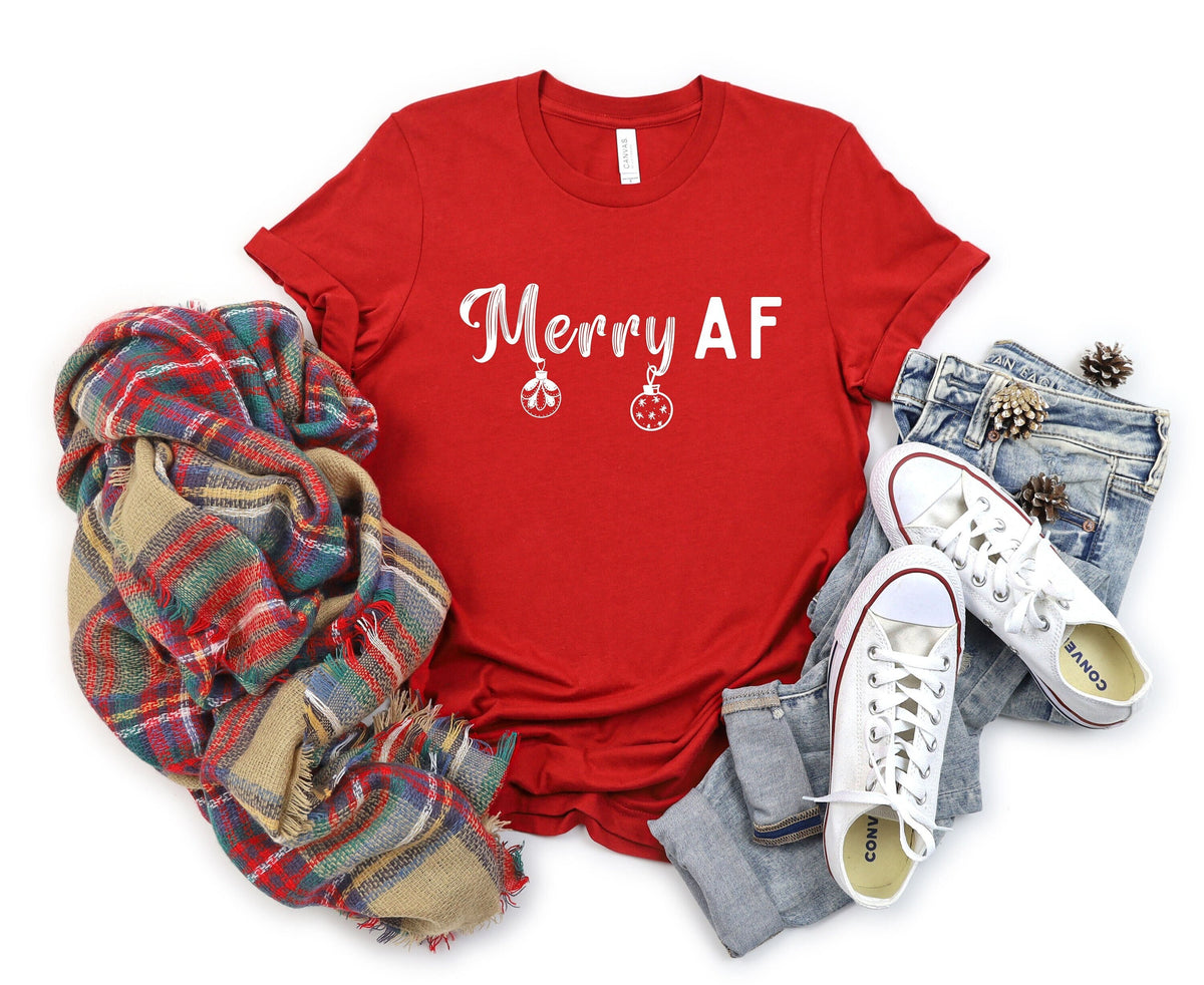 Merry AF T-Shirt in red laid flat from above beside a plaid scarf, jeans, sneakers and pinecones.