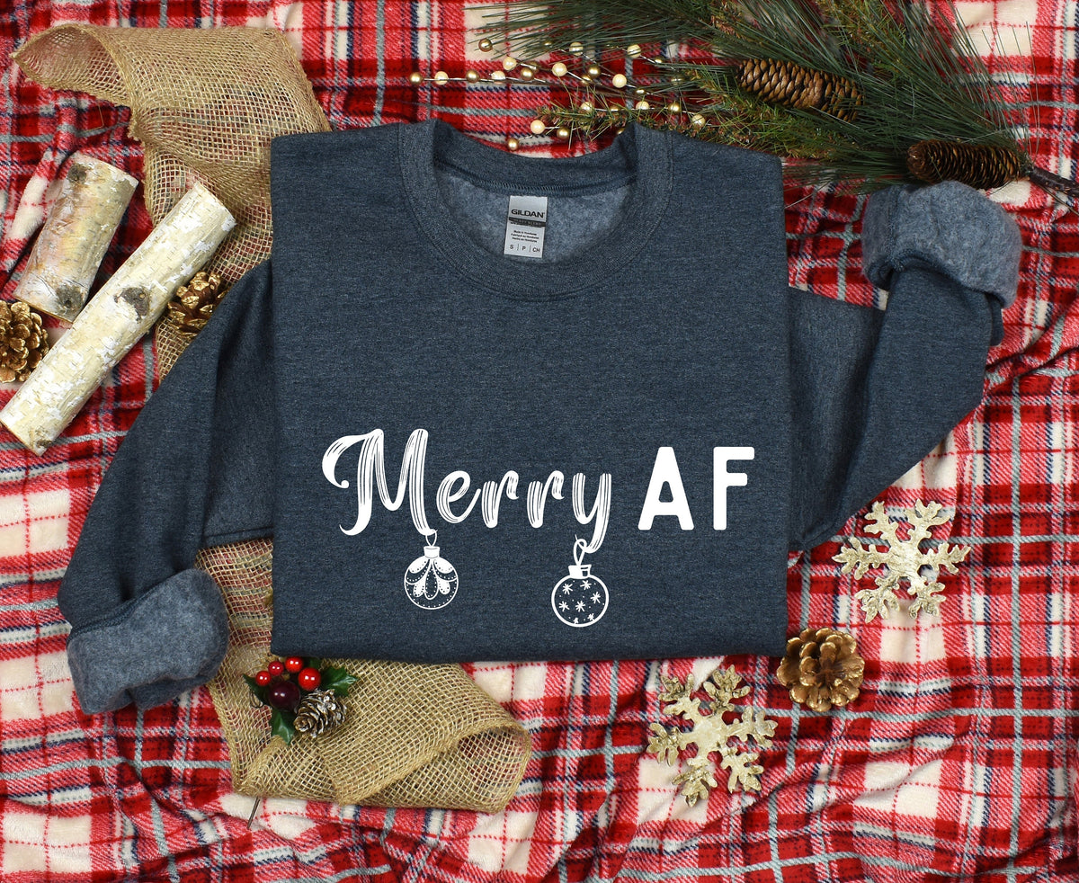 Merry AF Christmas Sweatshirt in Dark Heather flat-laid on red plaid surrounded by pine, cones, birch and gold snowflakes.